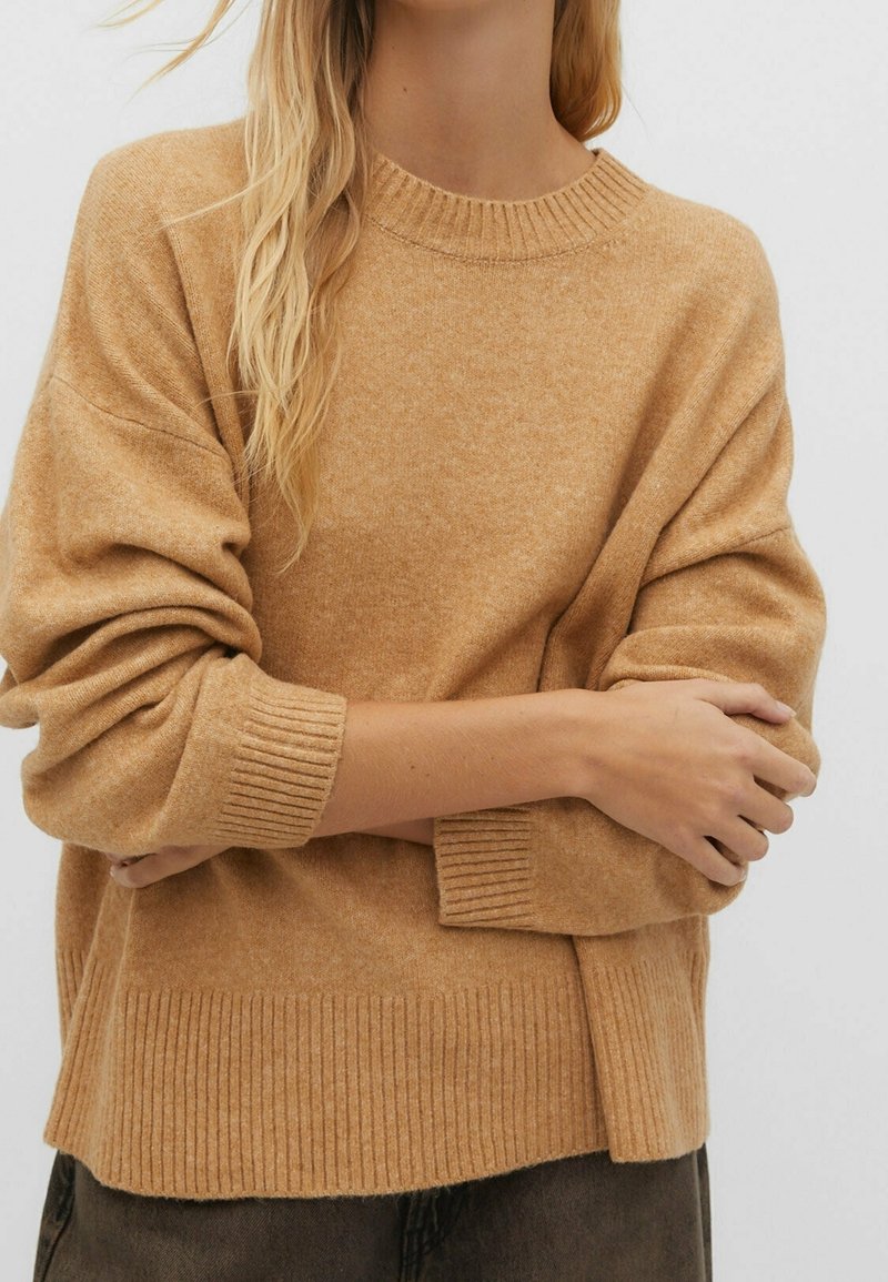 Personne blonde portant un pull tricoté oversize de couleur camel avec des poignets et un ourlet côtelés, les bras croisés sur la poitrine.