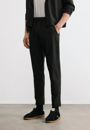 Muž v černých teplákových kalhotách, černé bundě a černých teniskách s gumovou podrážkou stojí s jednou rukou v kapse.