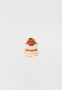 Sneaker Lacoste con corpo bianco, dettagli arancioni sul tallone e sulla suola, e un pannello texturizzato color tan sul retro.