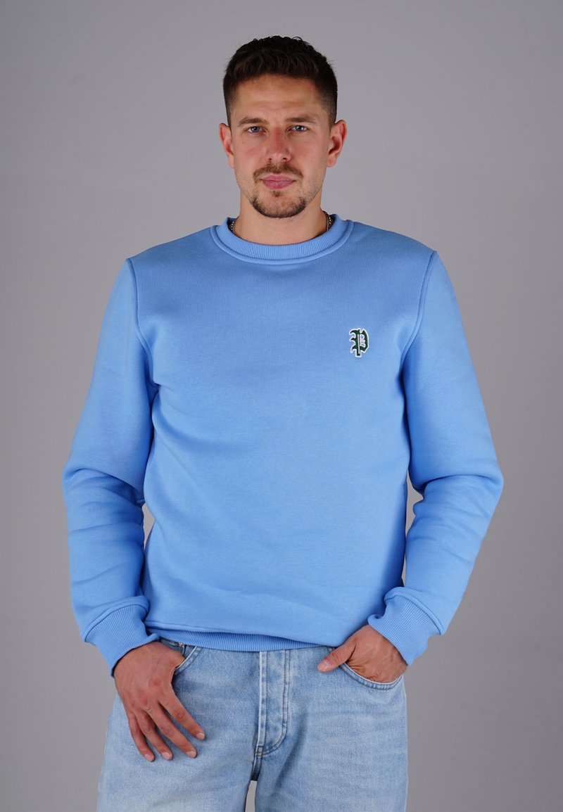 Hellblaues Sweatshirt mit Rundhalsausschnitt, gerippten Bündchen und einem kleinen grünen gestickten Logo auf der linken Brust, getragen mit heller Denim.