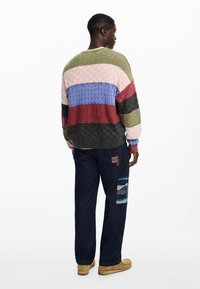 Multicolored knit sweater with textured pattern, featuring shades of green, pink, blue, and maroon. Paired with dark denim pants and beige footwear.