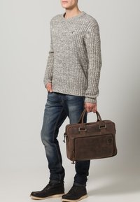 Bruine leren aktetas met dubbele handvatten, ritssluiting en een voorkoffer. Geribbelde grijze trui en blauwe jeans gecombineerd met zwarte laarzen.