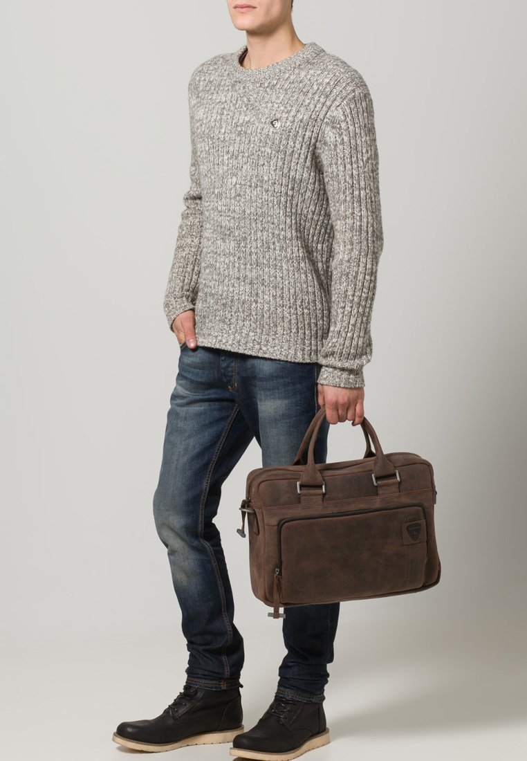 Bruine leren aktetas met dubbele handvatten, ritssluiting en een voorkoffer. Geribbelde grijze trui en blauwe jeans gecombineerd met zwarte laarzen.