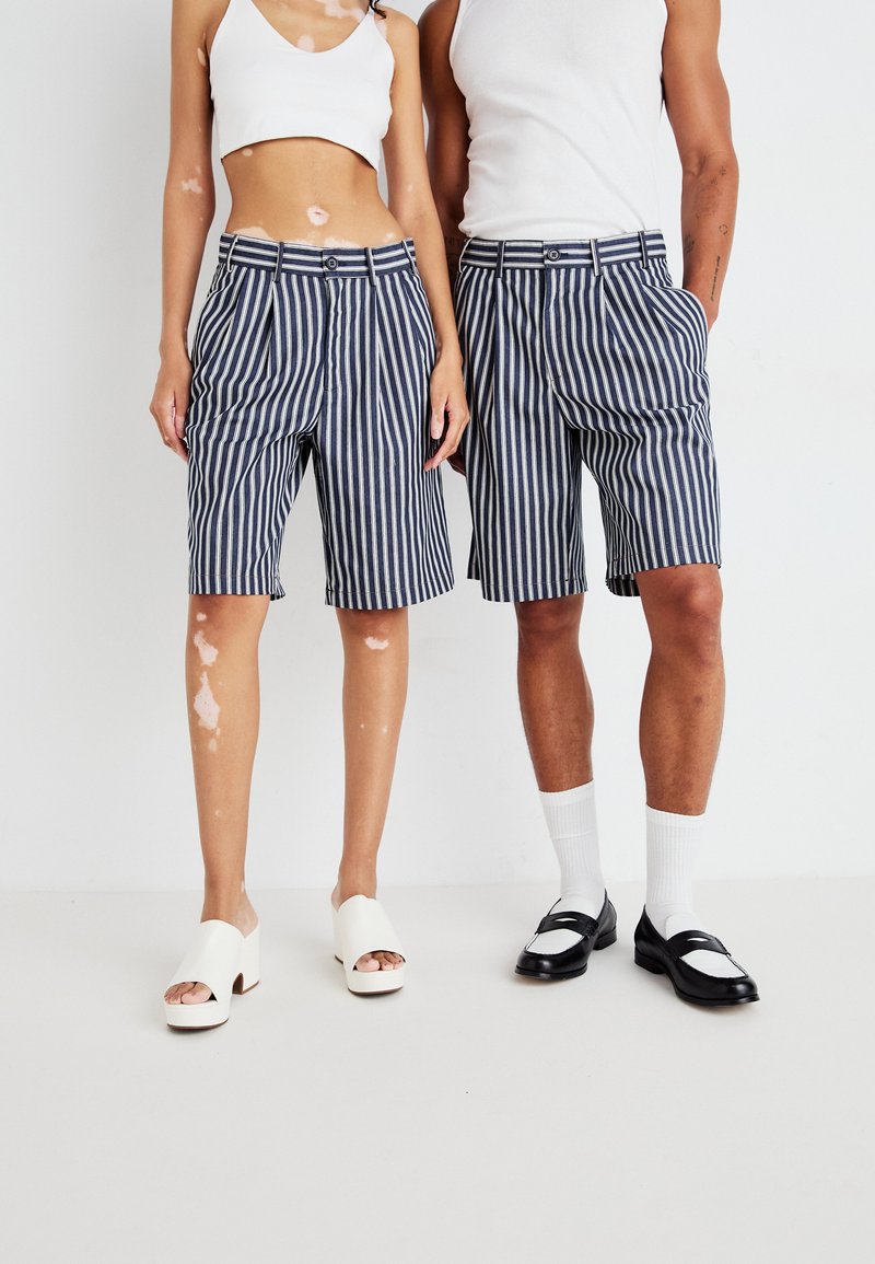 Stripete blå og hvite shorts med folder, kombinert med en hvit crop topp og blokkhelede mules. Skoene og sokkene kontrasterer i stil.