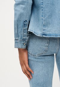 Χέρι ακουμπισμένο στο ισχίο, φορώντας ανοιχτό μπλε τζιν μπουφάν και τζιν παντελόνι που ταιριάζουν, με ορατή πίσω τσέπη και κουμπί στο μανίκι.