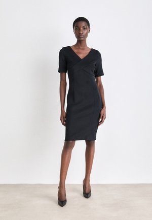 Black fitted dress with short sleeves, V-neck, and cross-over bodice. Smooth texture and knee-length cut. Worn with black high heels.