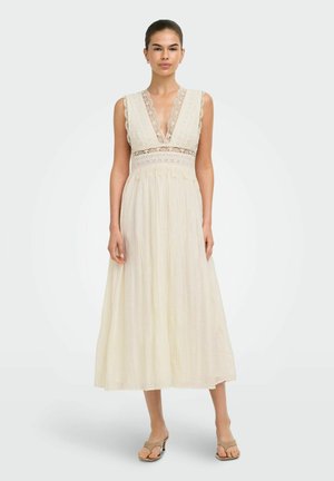 Mujer que lleva un vestido midi sin mangas en color crema con detalles de encaje y un escote en V profundo, combinado con sandalias beige de punta abierta.
