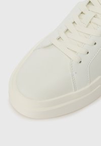 Bílé kožené tenisky s zaoblenou špičkou, širokou gumovou podrážkou a plochými tkaničkami. Jemné švy podél stran.