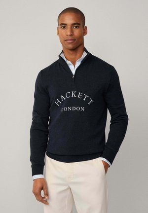 Maglione blu navy con colletto alto, design a mezza zip e logo bianco "HACKETT LONDON" ben visibile sul petto. Texture liscia e vestibilità aderente.