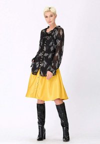 Blouse noire transparente avec motif floral et boutons, associée à une jupe jaune à longueur genou avec des détails en boutons. Le modèle porte des bottes noires haute jusqu'au genou.