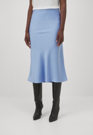 Femme portant une jupe midi bleu clair à ourlet évasé, associée à des bottes noires à talons montant jusqu'au genou et un haut blanc.