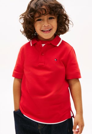 Copil zâmbitor cu păr creț, purtând un tricou polo roșu și blugi închiși la culoare, stând cu o mână în buzunar, pe un fundal alb.
