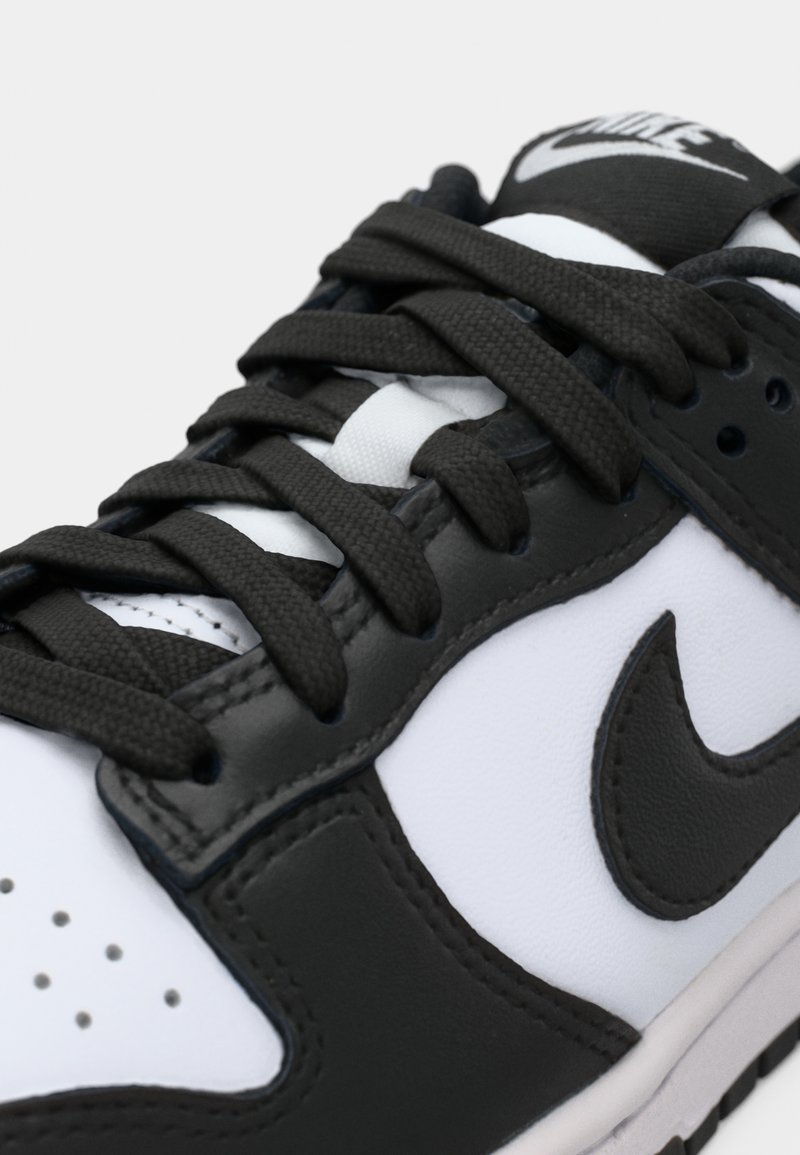 Zapatilla Nike en blanco y negro que presenta materiales de cuero y textil, logo prominente, sistema de cordones intrincado y puntera perforada para ventilación.