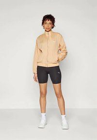 Ανοικτό ροδακινί ζακέτα με φερμουάρ και ψηλό γιακά, συνδυασμένη με μαύρες εφαρμοστές βερμούδες και λευκά αθλητικά παπούτσια. Απλός σχεδιασμός, χωρίς σχέδια.