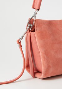 Sac à main en suédine corail, avec un accent en cuir lisse, doté d'un fermoir en métal et d'une bandoulière ajustable. Le sac présente une forme douce et structurée.
