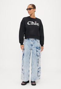 Μαύρο κοντό φούτερ με το "Chic" τυπωμένο στα λευκά, συνδυασμένο με υπερμεγέθεις ανοιχτό μπλε τζην cargo παντελόνες. Το μοντέλο φοράει σκούρα γυαλιά ηλίου και χοντρές παπούτσια.