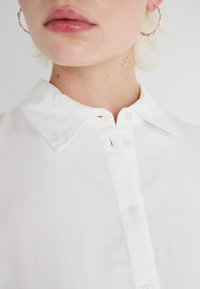 Chemise blanche avec un col à bouton, présente un léger éclat, une texture lisse et deux boutons visibles au cou. Boucle d'oreille en argent en forme de cercle.