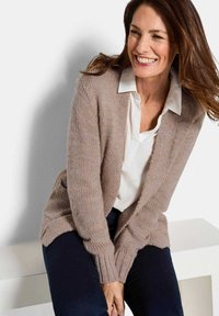 Lächelnde Frau mit langen braunen Haaren, die einen beigen Strickcardigan, eine weiße Bluse und dunkle Hosen trägt, sitzt auf einer weißen Bank vor einem schlichten Hintergrund.