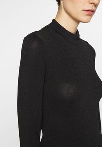 Haut à col roulé noir en tissu texturé et scintillant. Présente une silhouette ajustée et des manches longues, mettant en valeur un design chic et moderne.