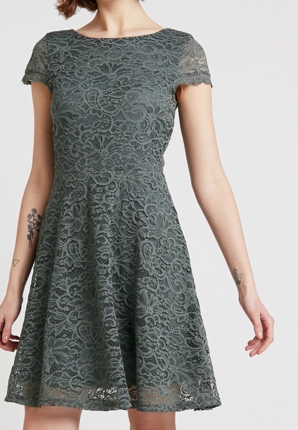 Femme portant une robe en dentelle gris foncé à manches courtes avec un motif floral et une taille cintrée, montrant ses avant-bras tatoués sur un fond uni.