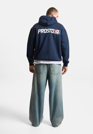 Osoba ubrana w granatową bluzę z kapturem "PROSTO." i jasnoniebieskie szerokie dżinsy, stojąca tyłem na jednolitym białym tle.