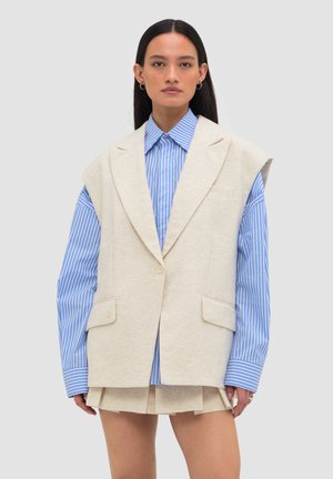 Frau trägt einen übergroßen beigen ärmellosen Blazer über einem blau-weiß gestreiften Langarmhemd mit passendem beigen Rock vor einfarbigem Hintergrund.