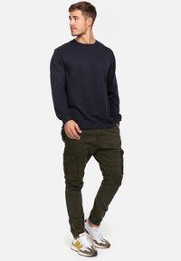 Donkerblauwe sweatshirt, ronde hals, lange mouwen; olijf groene cargobroek met zakken; witte en grijze sneakers met gele accenten.