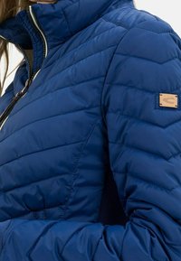 Tummansininen puhvitakki, jossa on tikattu muotoilu, stand-collari ja vetoketju. Hihassa on nahkakoriste, jossa on logo.