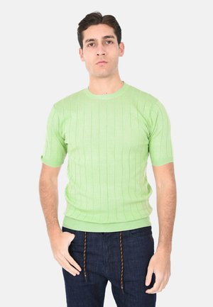 Uomo che indossa un maglione a maniche corte lavorato a maglia verde chiaro e jeans blu scuro, in piedi con una mano in tasca, su sfondo bianco.