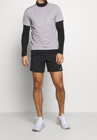 Camiseta atlética de manga corta gris con textura y mangas largas negras, combinada con pantalones cortos negros y zapatillas deportivas blancas, con detalles de logo.