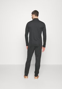 Sort langærmet skjorte og matchende bukser, lavet af glat stof, tætsiddende design, minimale sømme, båret med lysebrune sportsko.