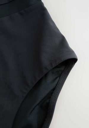 Close-up van zwarte stof en stiksels van een broekspijpopening van een hooggetailleerd kledingstuk tegen een lichte achtergrond.