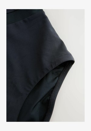 Close-up van zwarte stof en stiksels van een broekspijpopening van een hooggetailleerd kledingstuk tegen een lichte achtergrond.