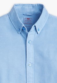 Chemise bleu clair à boutons avec une coupe décontractée. Confectionnée en tissu lisse, elle présente un col, des détails de couture et une broderie de logo subtile.