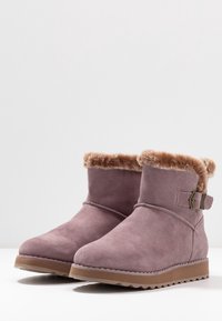 Fialové semišové kotníkové boty s umělou kožešinou na manžetě, bočním popruhem a texturovanou gumovou podešví. Jemný zaoblený tvar špičky.
