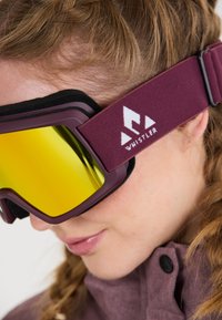 Skibril met een kastanjebruwe rand, geel-getinte lens en zwarte vulling. Beschikt over een merkriem met wit logo.
