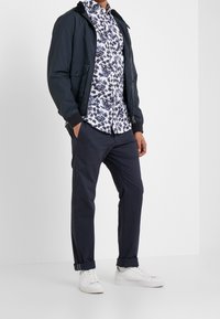 Giubbotto bomber della marina sopra una camicia bianca con motivo floreale blu, abbinato a pantaloni scuri e sneaker bianche. Tessuto morbido, vestibilità casual.