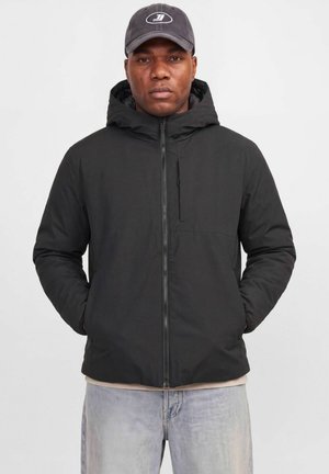 Un homme porte une veste noire à capuche et à fermeture éclair ainsi qu’une casquette avec un logo. Il se tient debout, les mains dans les poches, devant un fond clair et uni.