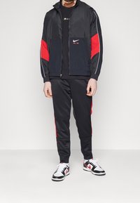 Nike träningsoverall bestående av en svart vindjacka med röda detaljer och dragkedja framtill, i kombination med svarta byxor och vita sneakers med röda inslag.