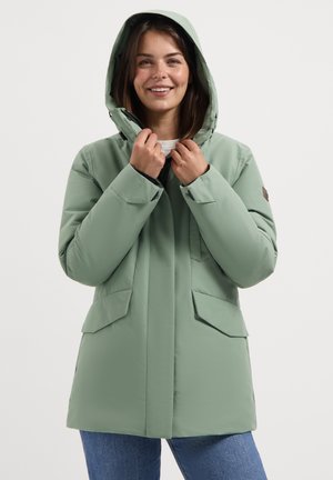 Vrouw die glimlacht, gekleed in een lichtgroene winterjas met capuchon en voorzakken, met blauwe jeans, en de kraag van de jas met beide handen vasthoudt.