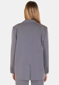 Blazer grigio su misura con revers a lingua, due bottoni sui polsini e uno spacco sul retro. Tessuto liscio e strutturato.