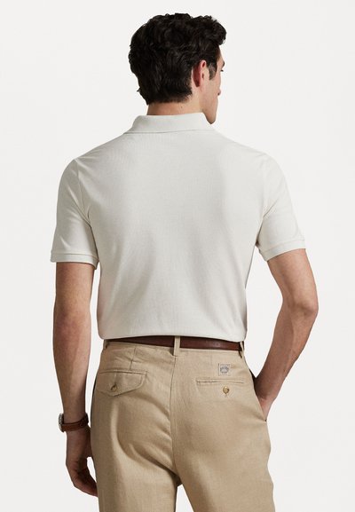 Πόλο με κοντά μανίκια σε εκρού χρώμα, με γιακά. Μπεζ παντελόνι με πίσω τσέπες, διακοσμημένο με λογότυπο, και καφέ δερμάτινη ζώνη.