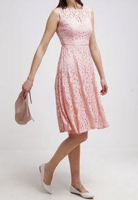 Růžové krajkové šaty s přiléhavým tělem, sukní po kolena a bez rukávů. Kombinováno s světle zbarvenými balerínami a malou kabelkou.