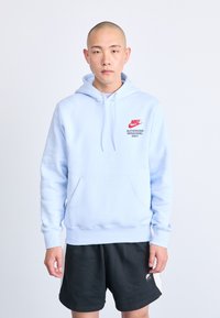 Šviesiai mėlyna Nike džemperis su priekiu kišene, su raišteliu reguliuojama kapucha ir raudonais užrašais "TIK ĮGALIOTAM PERSONALUI" ant krūtinės. Juodos šortai.