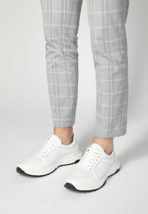 White athletic sneakers with a textured upper, featuring mesh panels and black rubber soles, paired with gray plaid trousers.