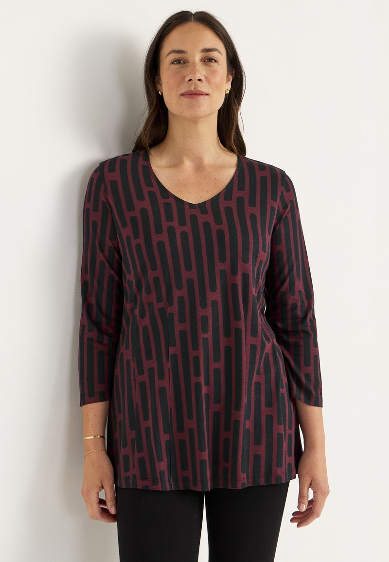 Vrouw in een bordeauxrode lange mouwen top met zwart rechthoekig patroon, zwarte broek, staande tegen een witte muur.