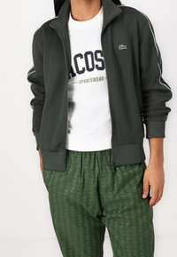 Chaqueta verde oscura con cremallera y detalles en blanco, combinada con una camiseta blanca con un logo, y pantalones verdes con patrón y cintura elástica.