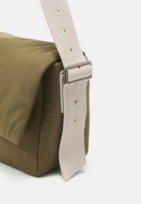 Bolso cruzado color caqui con un exterior de tela suave. Presenta una amplia correa de cuero beige y herrajes metálicos plateados para ajustes.