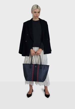 Borsa a mano blu scuro con motivo a diamante, caratterizzata da una striscia rossa centrale e manici superiori. Il modello indossa un cappotto nero sopra un maglione grigio.