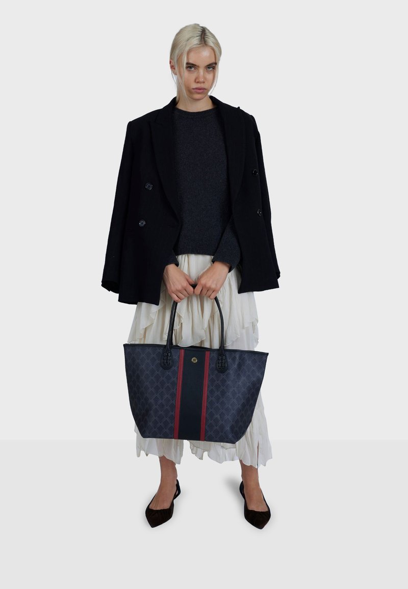 Platnena torba tamnoplave boje s dijamantnim uzorkom, središnjim crvenim prugom i gornjim ručkama. Model nosi crni kaput preko sive veste.